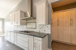 Modern kitchen featuring white cabinetry, dark stone countertops, a gas cooktop with a stainless steel range hood, mosaic tile backsplash, and extensive pantry storage