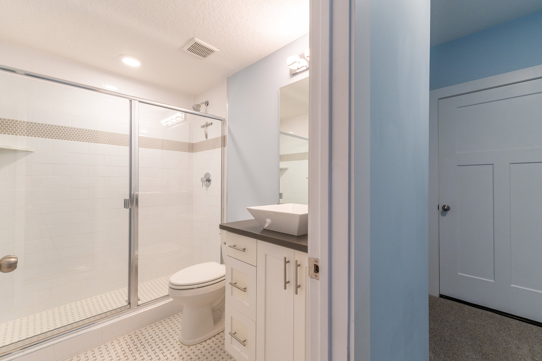 Modern bathroom featuring a sliding glass shower enclosure, white subway tile with a decorative accent band, and a white floating vanity with a vessel sink