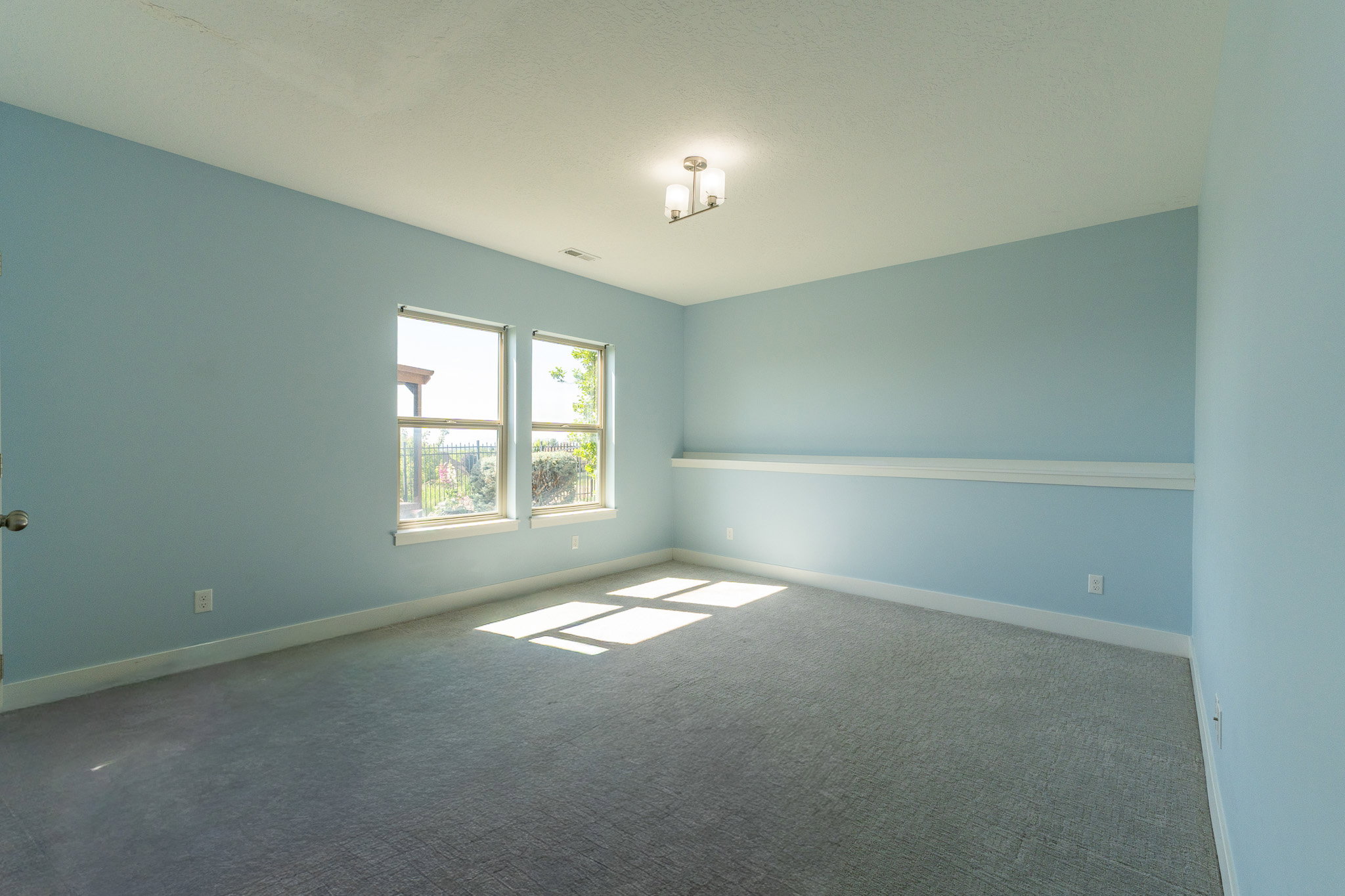 Bright room featuring two windows, light blue walls, gray textured carpet, white baseboards, and a contemporary ceiling fixture