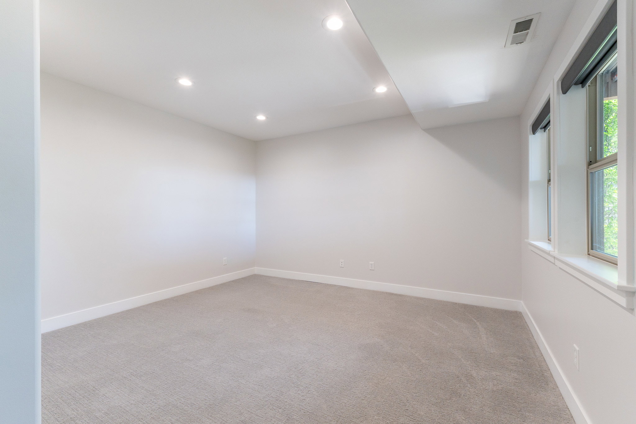 Spacious room featuring light gray carpeting, recessed ceiling lighting, and multiple windows with roller shades