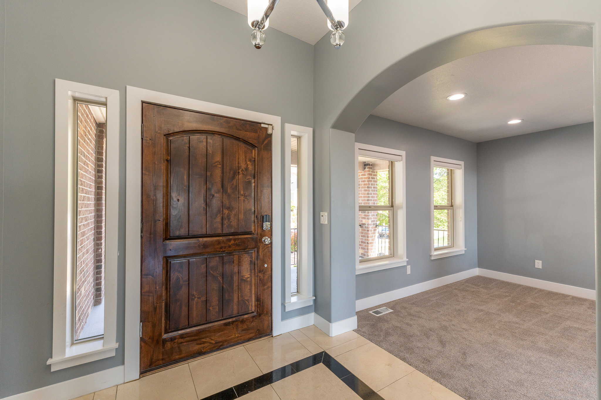 Grand entry featuring a paneled wood door with sidelight windows, tile flooring with a dark accent border, an overhead chandelier, and an arched entryway to a separate room with carpet flooring and windows