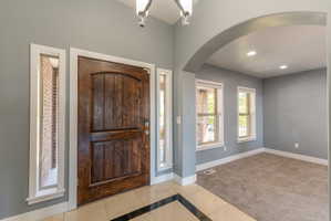Grand entry featuring a paneled wood door with sidelight windows, tile flooring with a dark accent border, an overhead chandelier, and an arched entryway to a separate room with carpet flooring and windows