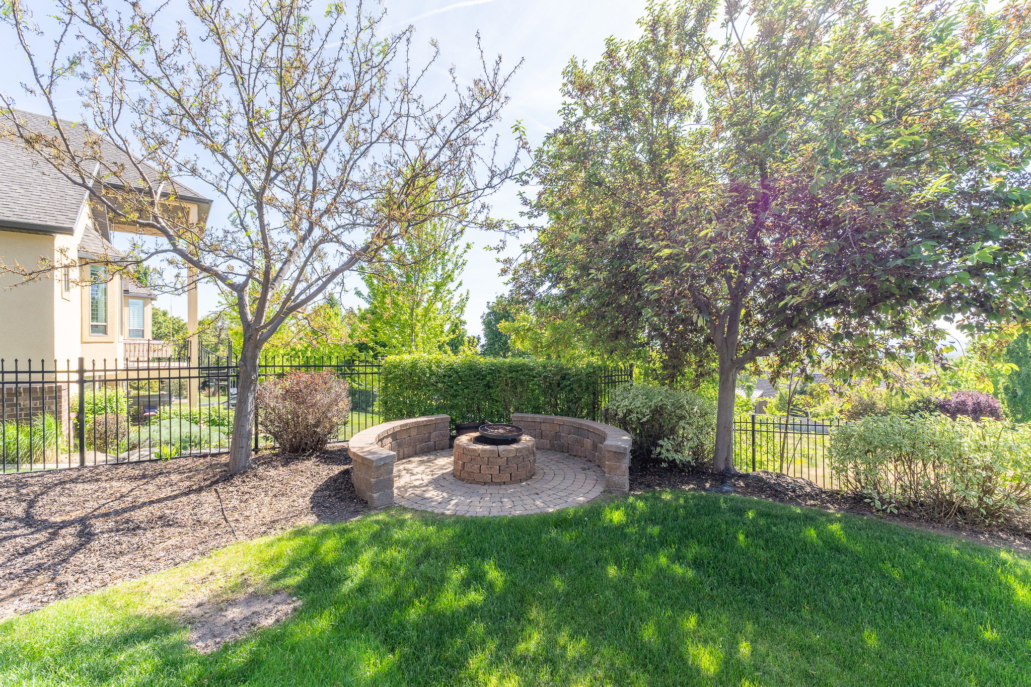 Outdoor fire pit with a brick-paver base and curved stone retaining walls, surrounded by mature trees and landscaping