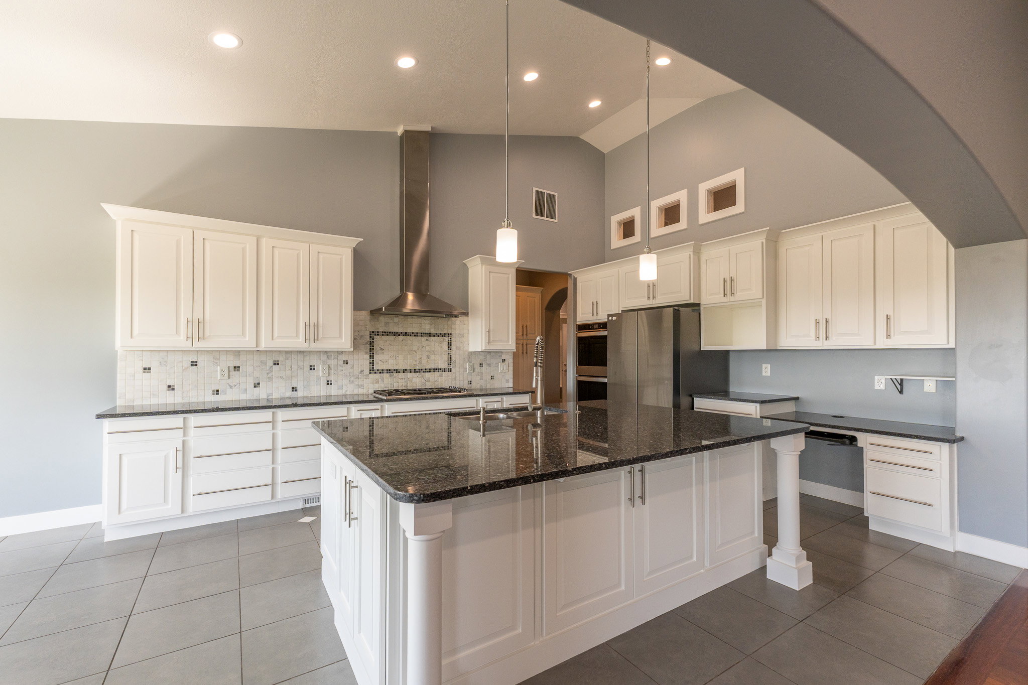 Vaulted ceiling kitchen featuring white cabinetry, dark stone countertops, stainless steel appliances, a center island with a built-in sink, and tile flooring