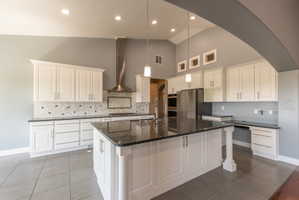 Vaulted ceiling kitchen featuring white cabinetry, dark stone countertops, stainless steel appliances, a center island with a built-in sink, and tile flooring
