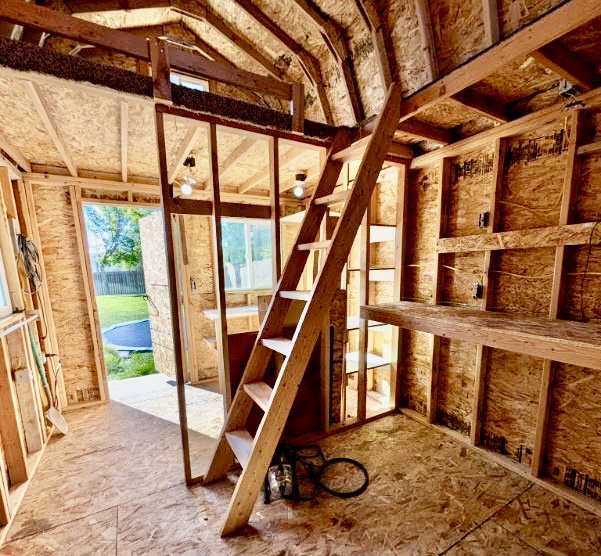 Unfinished interior space featuring exposed wood framing, oriented strand board walls and flooring, and a built-in wooden ladder providing access to an upper loft