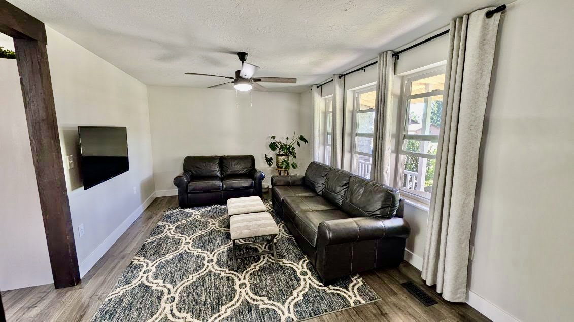 Living area featuring wood-finish flooring, white baseboards, a ceiling fan with integrated lighting, and multiple windows with light-colored drapes