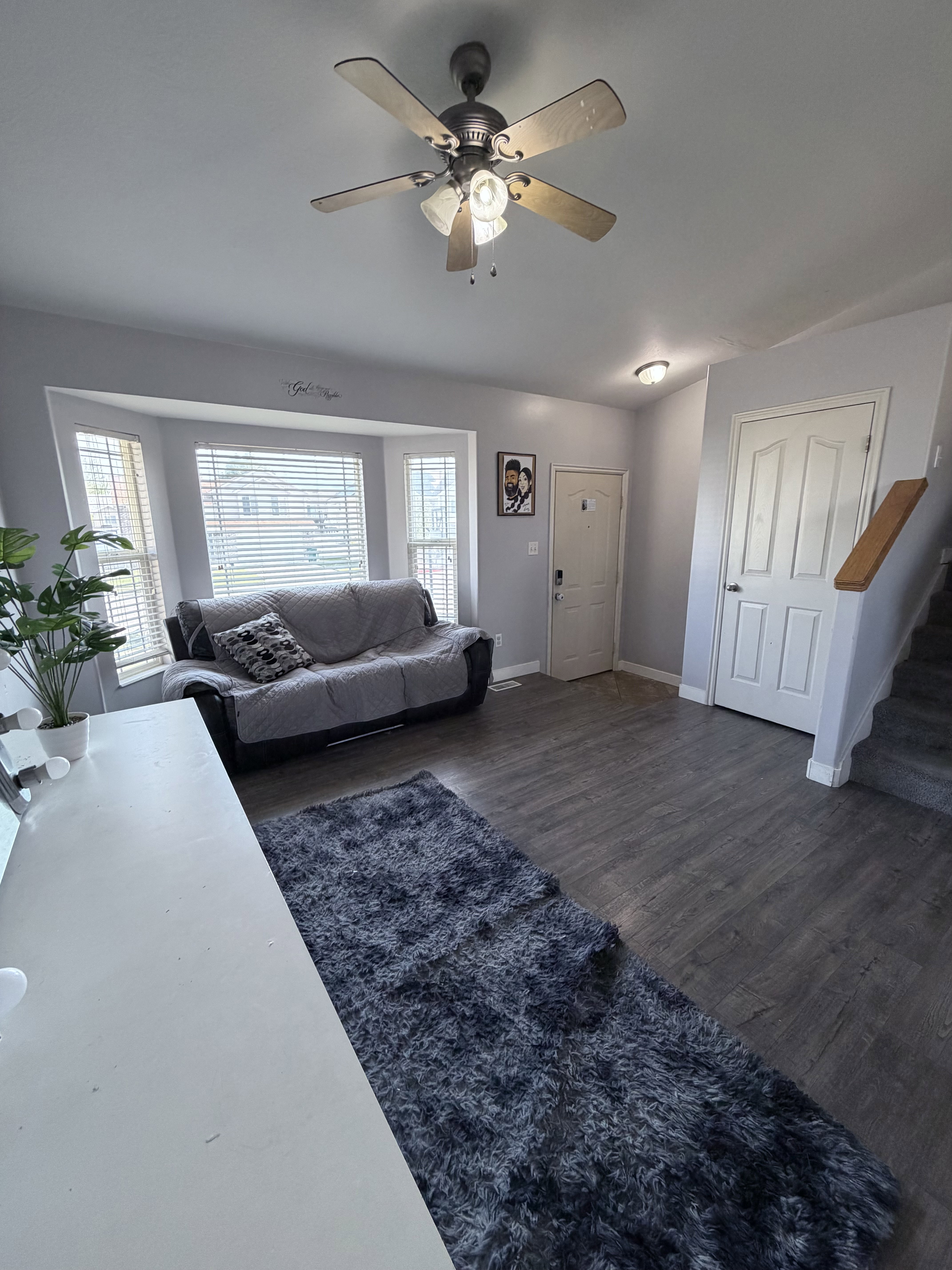 Spacious living area featuring wood-finish flooring, a ceiling fan with integrated lighting, and a bay window with blinds