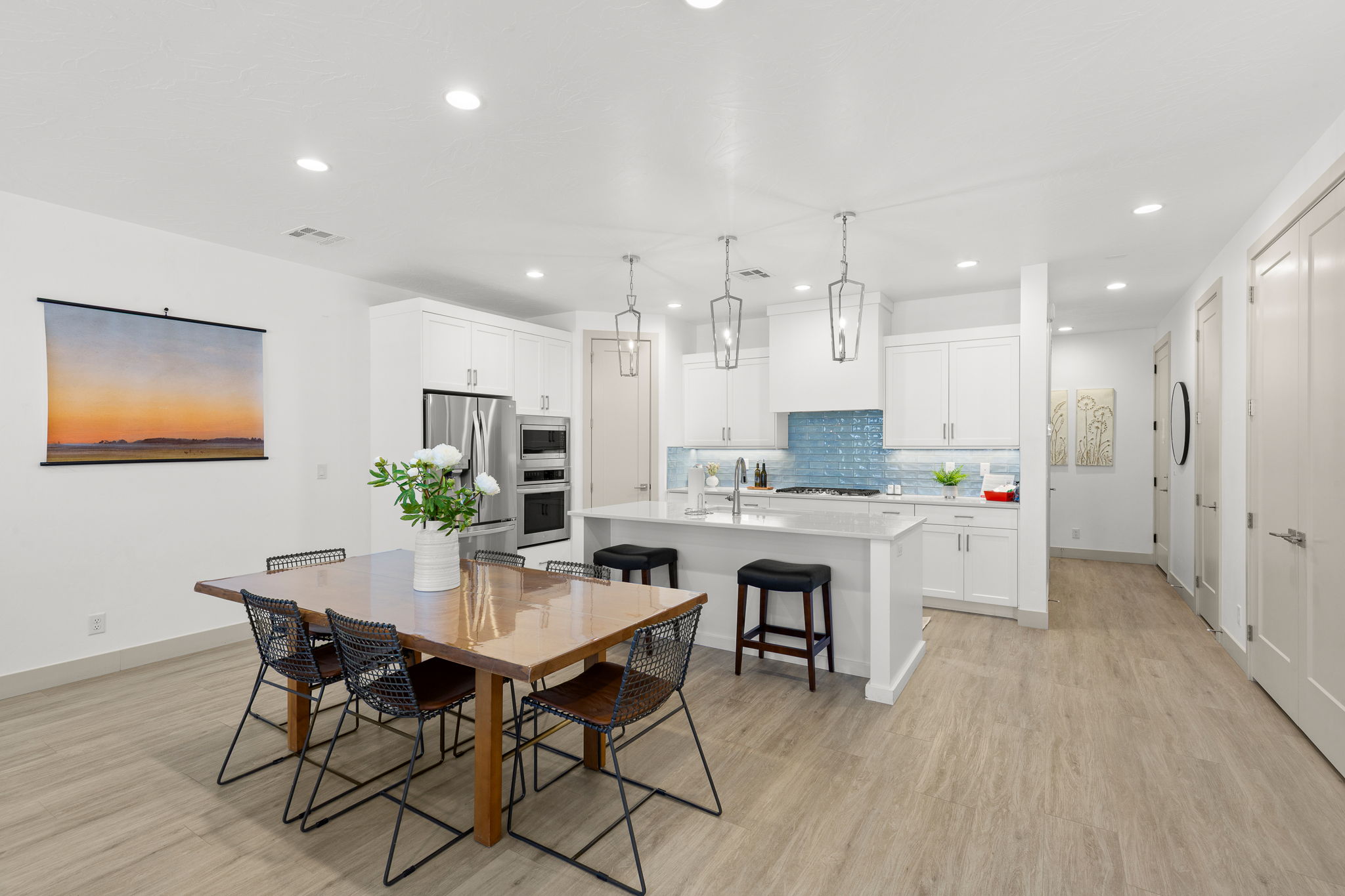Open-concept kitchen and dining area featuring light wood-finish flooring, white cabinetry with contemporary hardware, stainless steel appliances, a blue subway tile backsplash, and recessed lighting