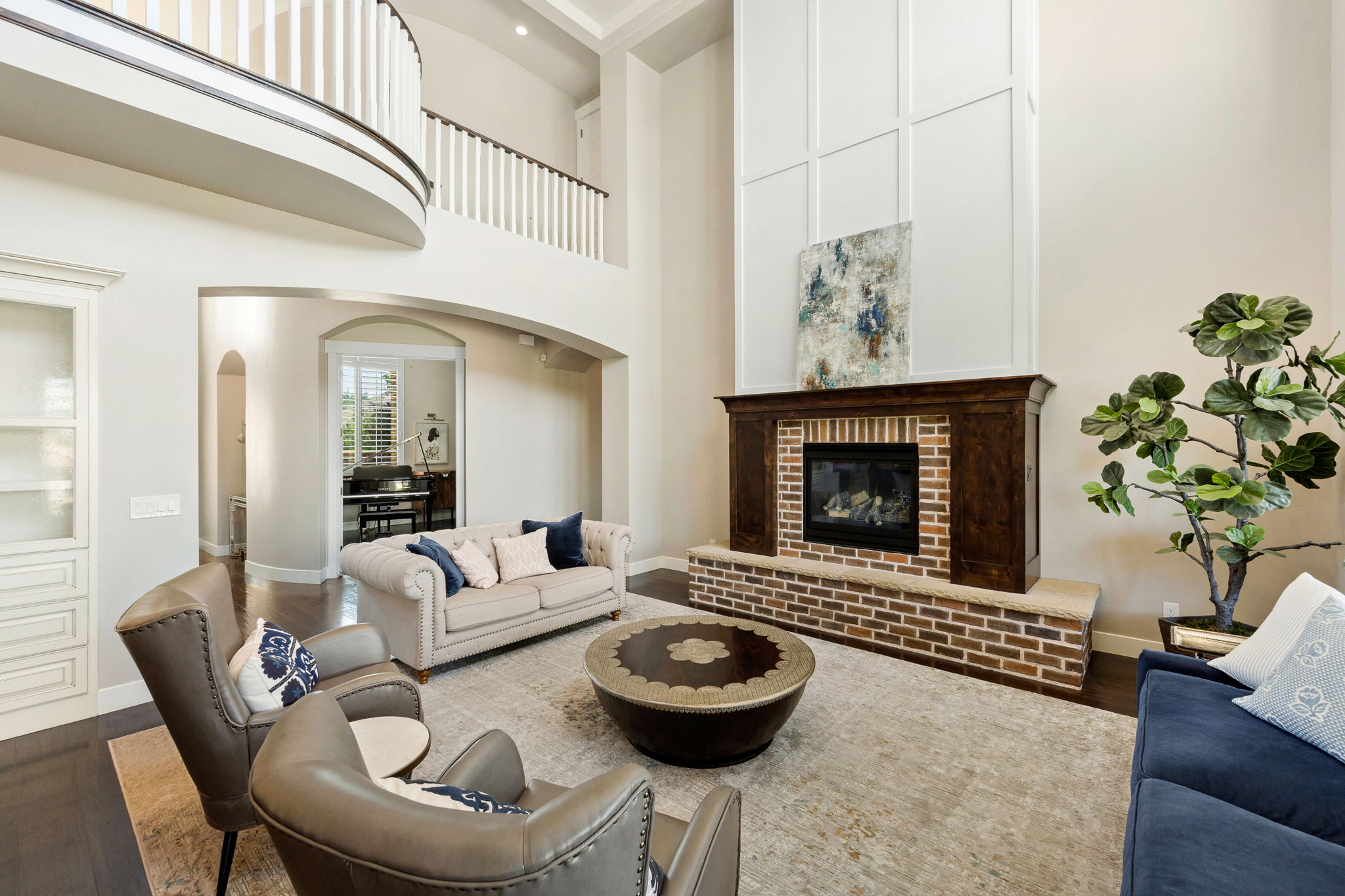 Two-story living area featuring a brick and wood-finish fireplace, rich wood-finish flooring, a curved balcony with white balusters, and an arched entryway