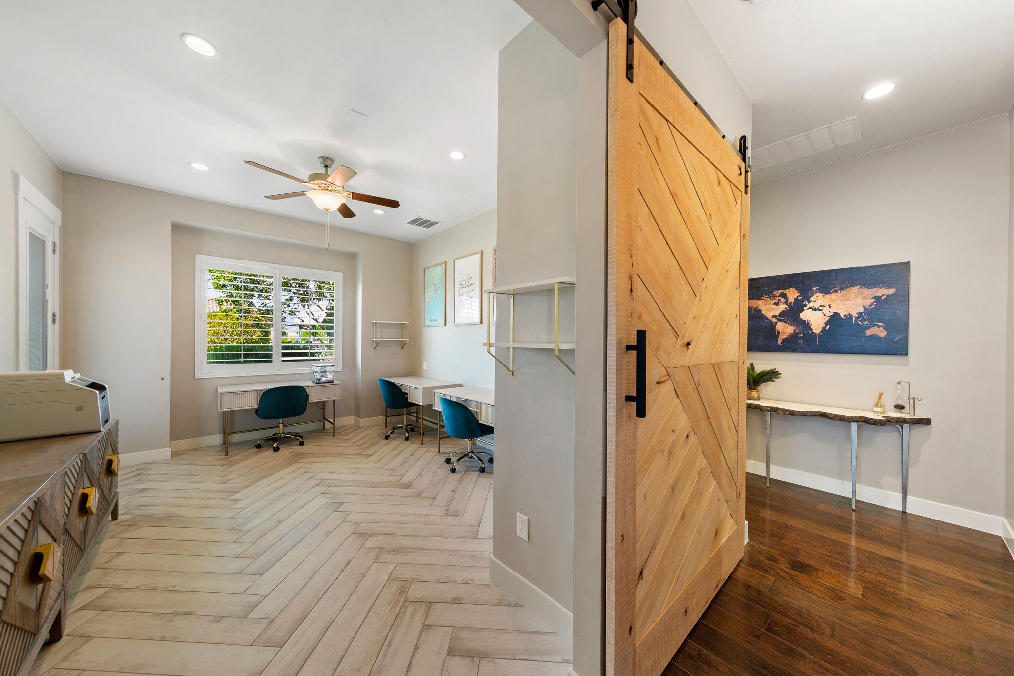 Natural wood barn door with black hardware opening to a room featuring light wood-finish flooring in a herringbone pattern, recessed lighting, a ceiling fan, and a window with plantation shutters