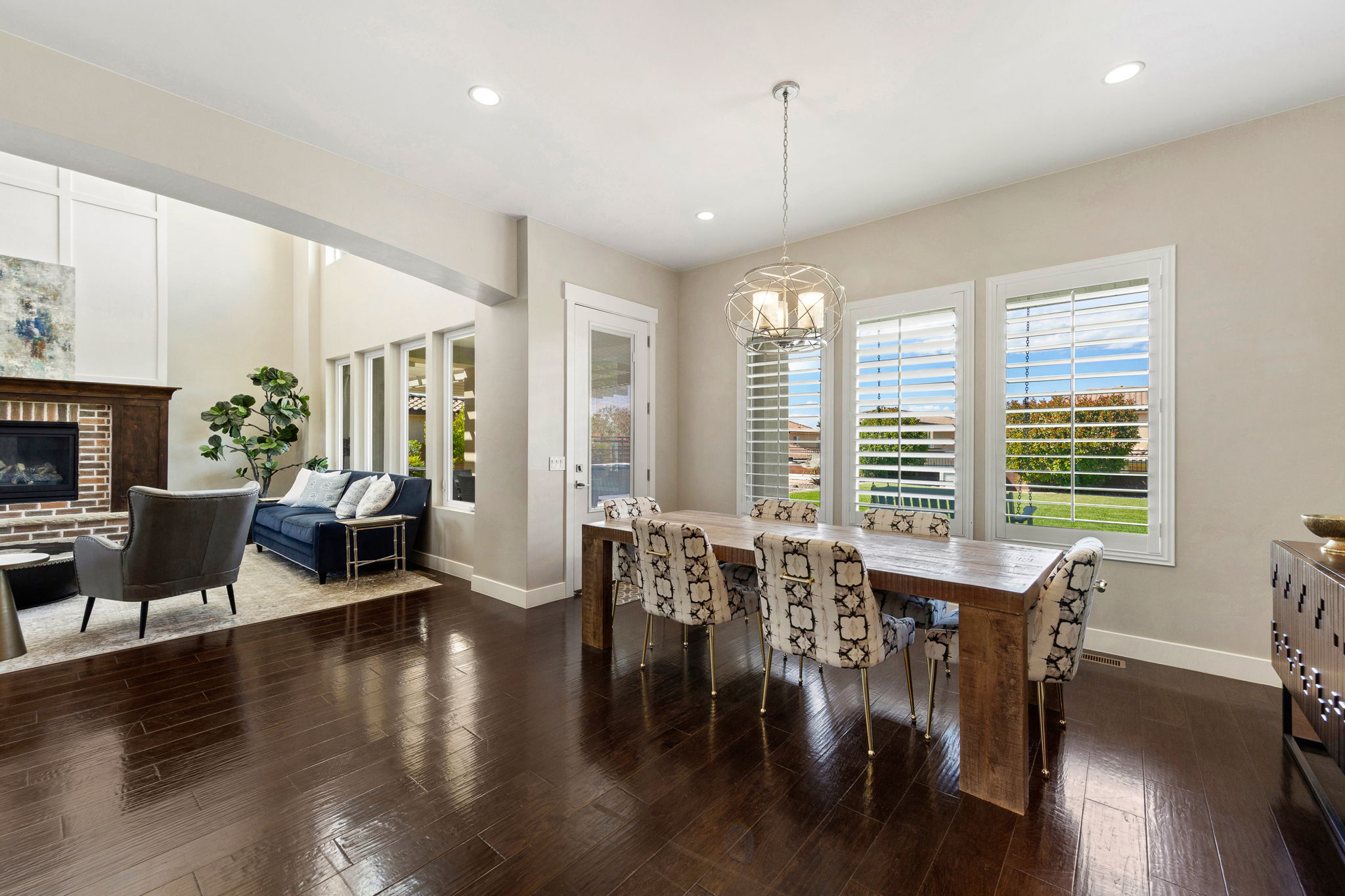 Open-concept dining area with dark wood-finish flooring, a contemporary chandelier, and three large windows featuring plantation shutters
