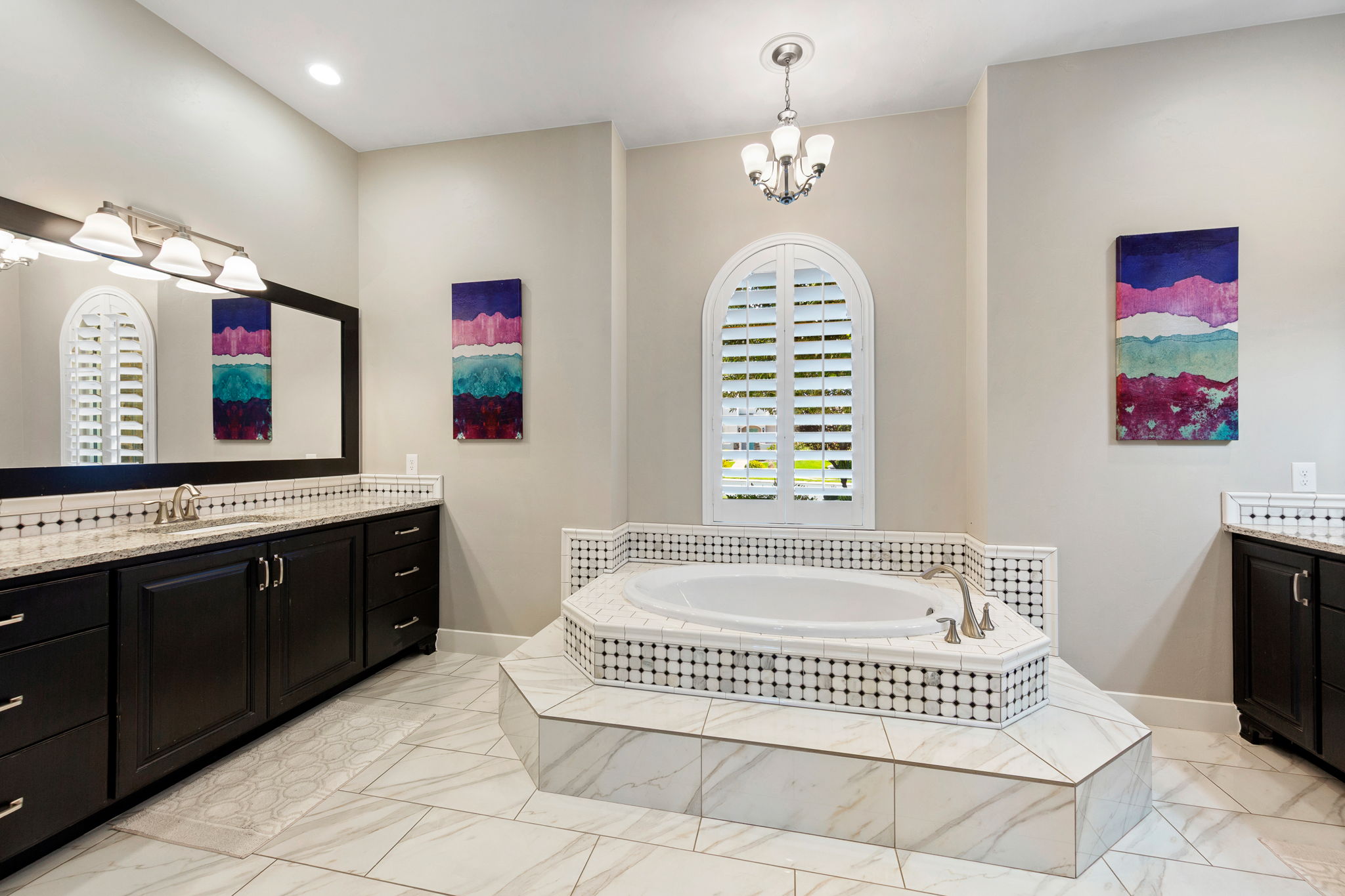 Spacious bathroom featuring a built-in corner soaking tub, arched window with plantation shutters, dual vanity with dark wood cabinetry, white countertops, and tile flooring