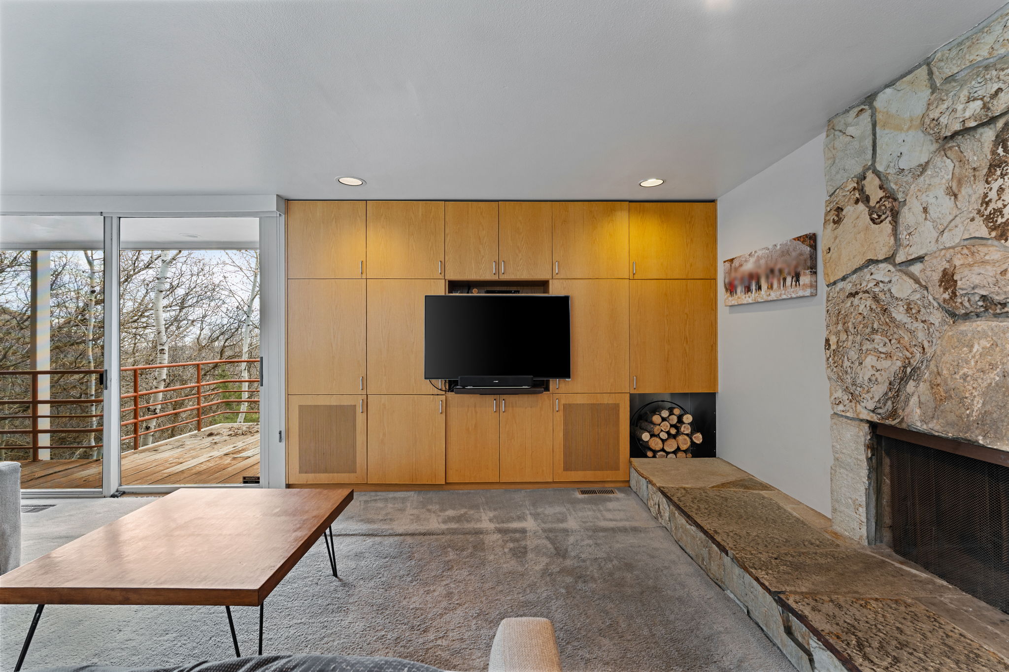 Living area featuring a floor-to-ceiling stone fireplace, built-in blonde wood cabinetry, recessed lighting, and sliding glass doors opening to an exterior deck