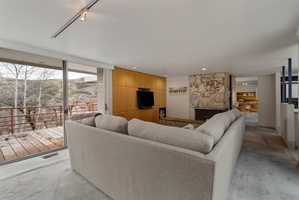 Spacious living area featuring a stone fireplace, wood-finish wall paneling with built-in media, track lighting, and sliding glass doors opening to an outdoor deck