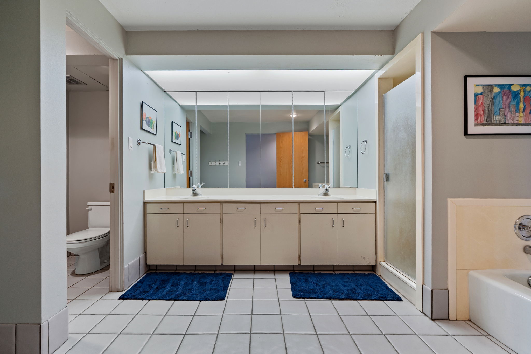 Spacious bathroom featuring a double vanity with ample counter space, extensive mirror cabinetry, a glass-enclosed shower, a separate commode area, and white tile flooring