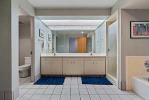 Spacious bathroom featuring a double vanity with ample counter space, extensive mirror cabinetry, a glass-enclosed shower, a separate commode area, and white tile flooring