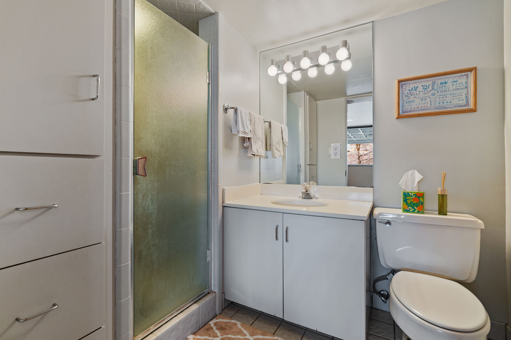 Bathroom featuring a vanity with an integrated sink, a rectangular mirror with a multi-bulb light fixture, a toilet, and a shower with a frosted glass door