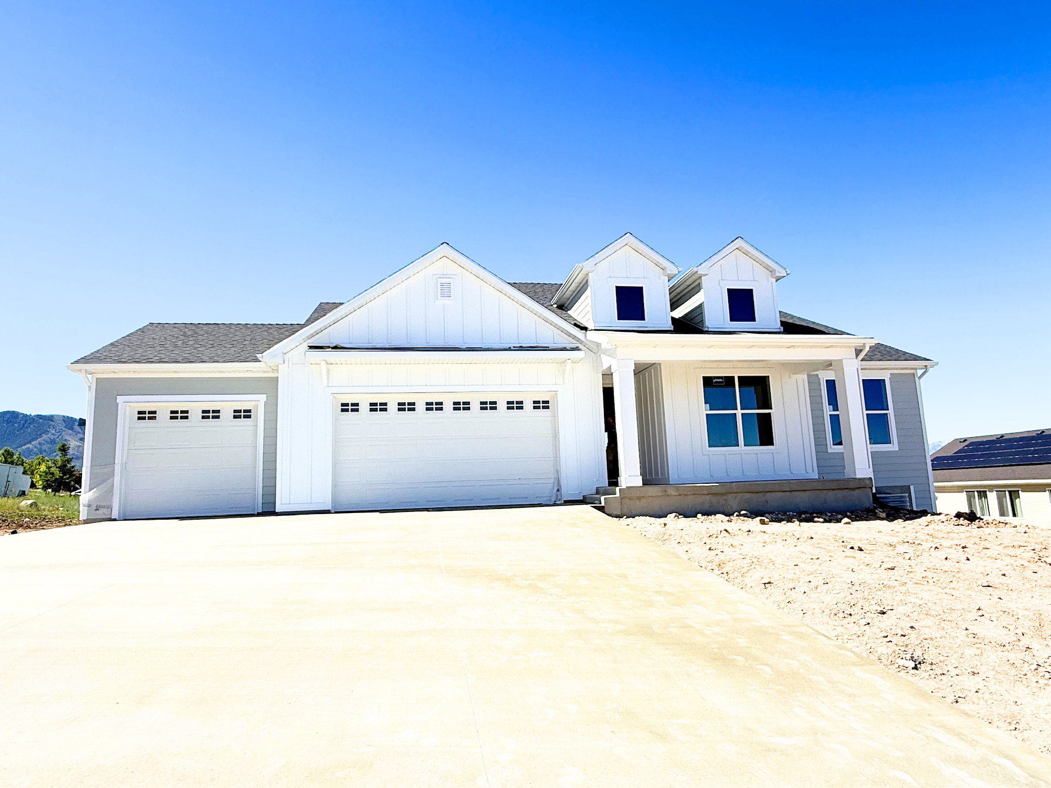 Contemporary ranch-style home featuring a light gray exterior, white trim, a covered entry porch with white columns, two dormer windows, and a multi-car garage with white doors