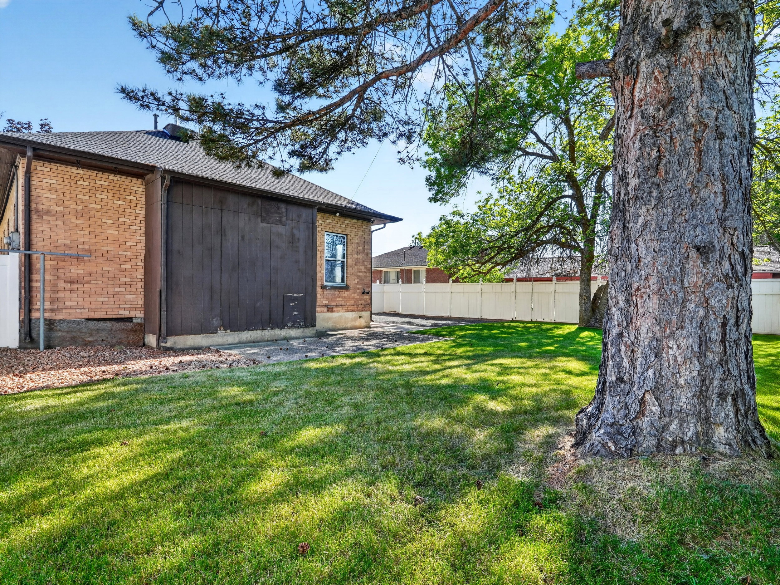Spacious exterior featuring a large tree, a manicured lawn, a white perimeter fence, and a brick-sided structure with a dark wood-paneled section