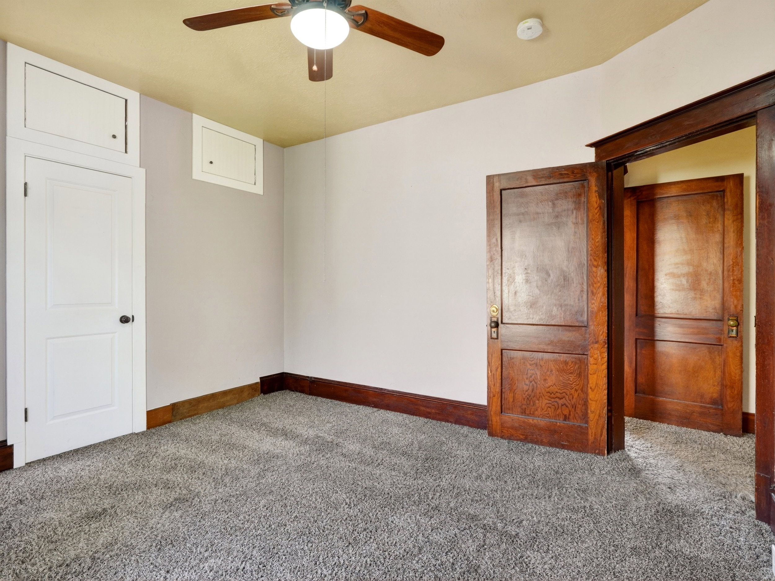 Carpeted room featuring a ceiling fan with integrated lighting, a white panel door with an overhead storage compartment, and two wood-finish doors with distinct wood grain