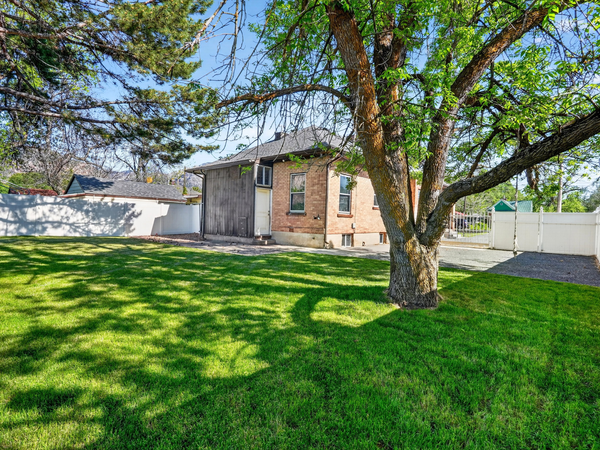 Expansive lawn area featuring a mature shade tree, a white privacy fence, a gravel driveway, and a brick exterior building with wood siding