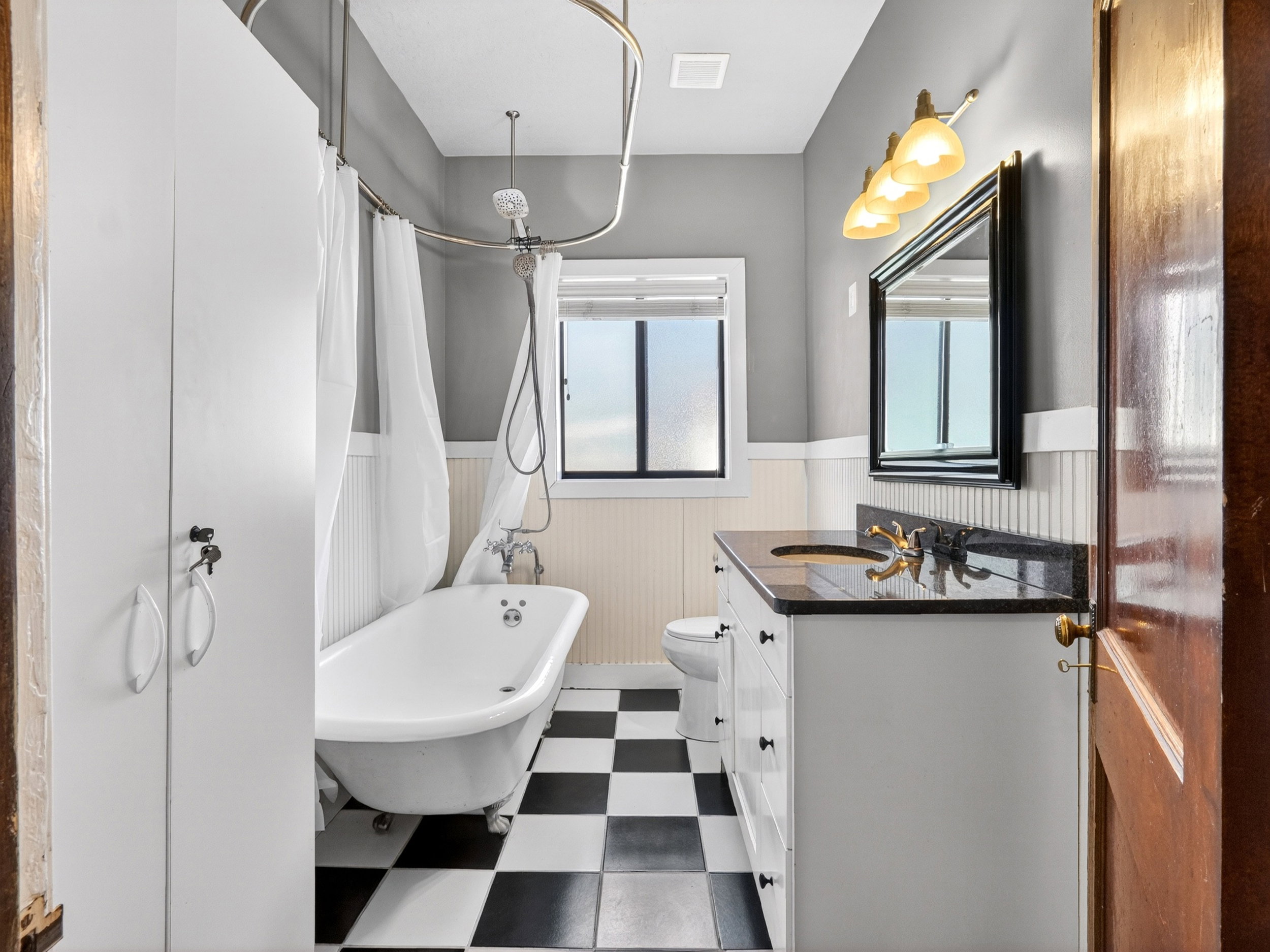 Bathroom featuring a clawfoot tub with shower enclosure, a vanity with dark countertop and undermount sink, a wall-mounted mirror with overhead lighting, and checkerboard floor tiles