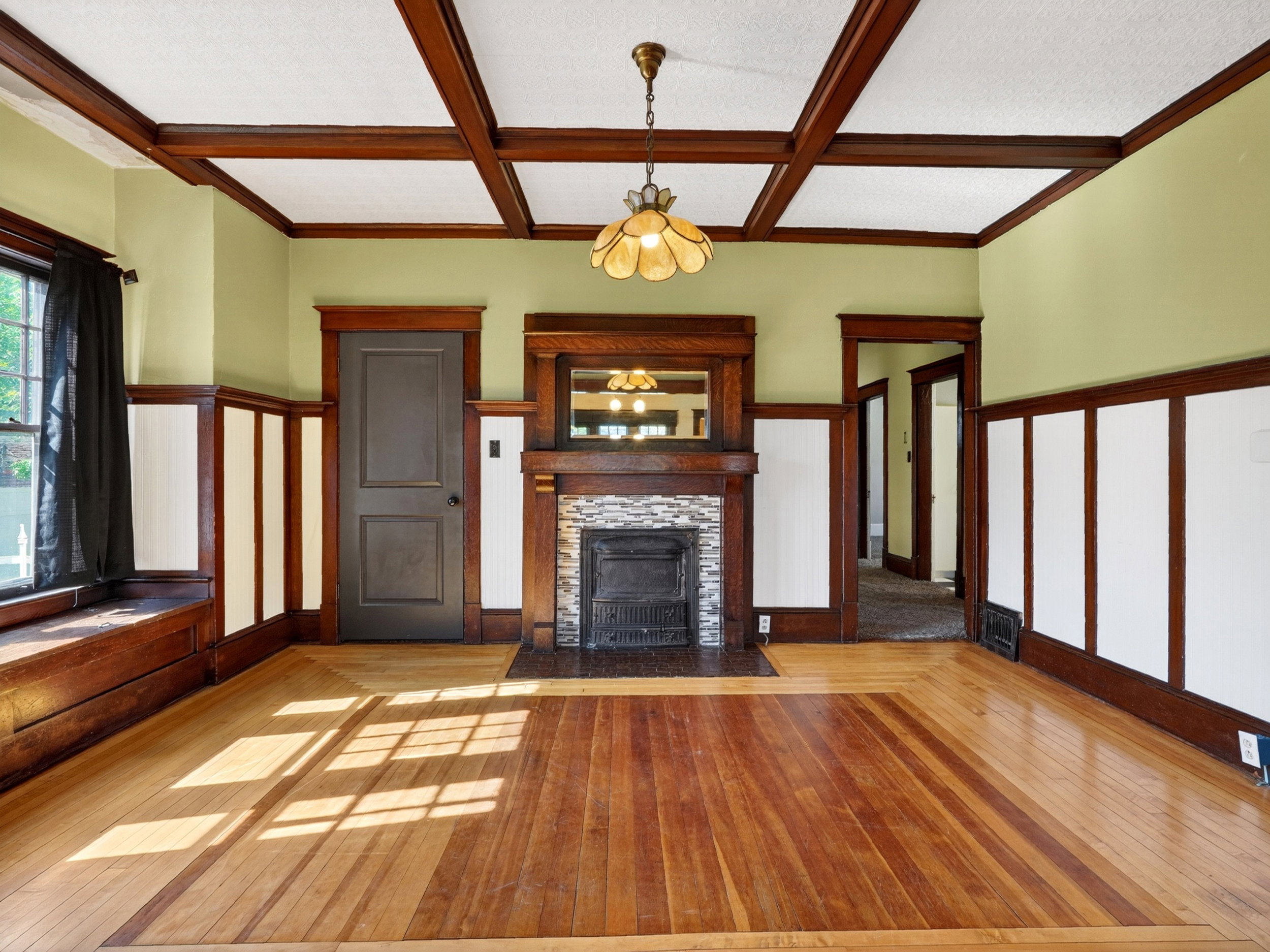 Hardwood flooring with border inlay, coffered ceiling, wood-paneled walls, built-in window bench, and a stone-clad fireplace with wood mantel