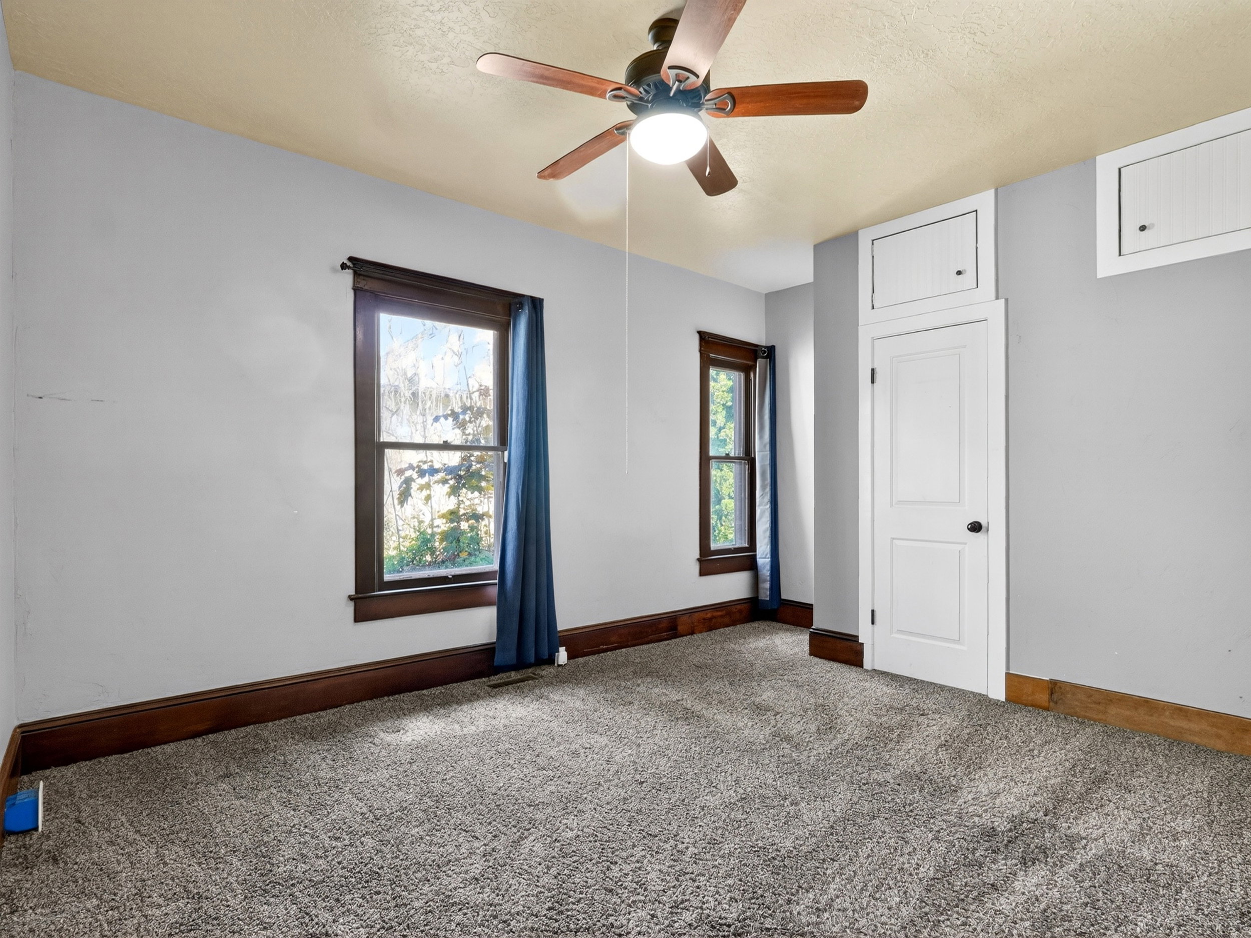 Neutral-toned carpeting grounds this space, complemented by a ceiling fan with light fixture and two windows with wood-trim framing