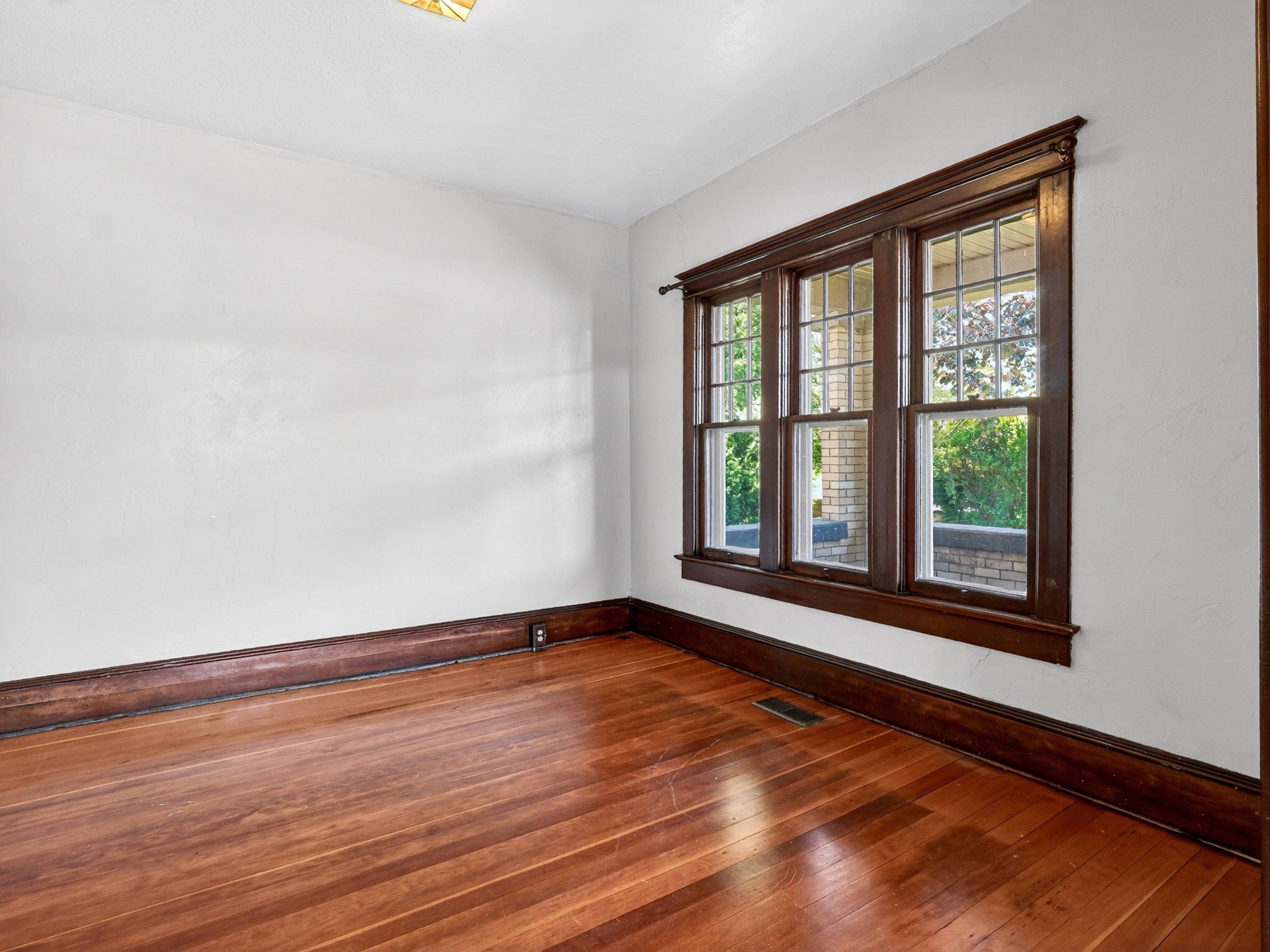 Room featuring rich wood-finish flooring, white walls, dark wood baseboards, and a triple window with detailed trim and muntins