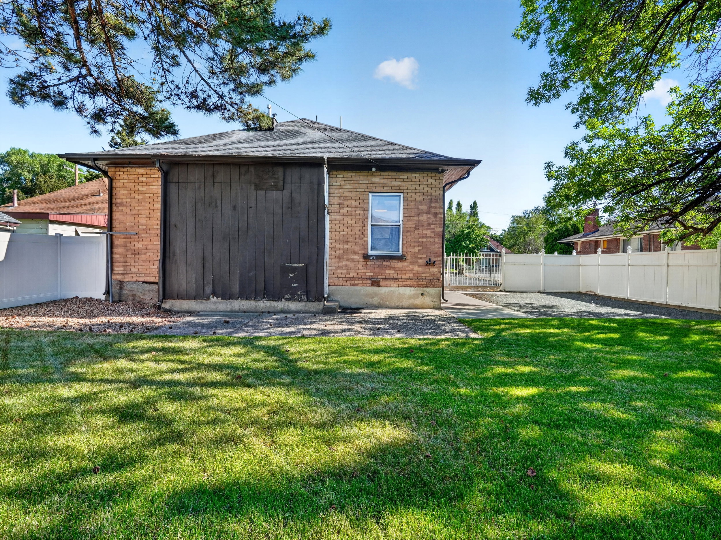 Expansive lawn area featuring a brick exterior structure, a dark wood-paneled wall, a white vinyl fence, and a gravel driveway