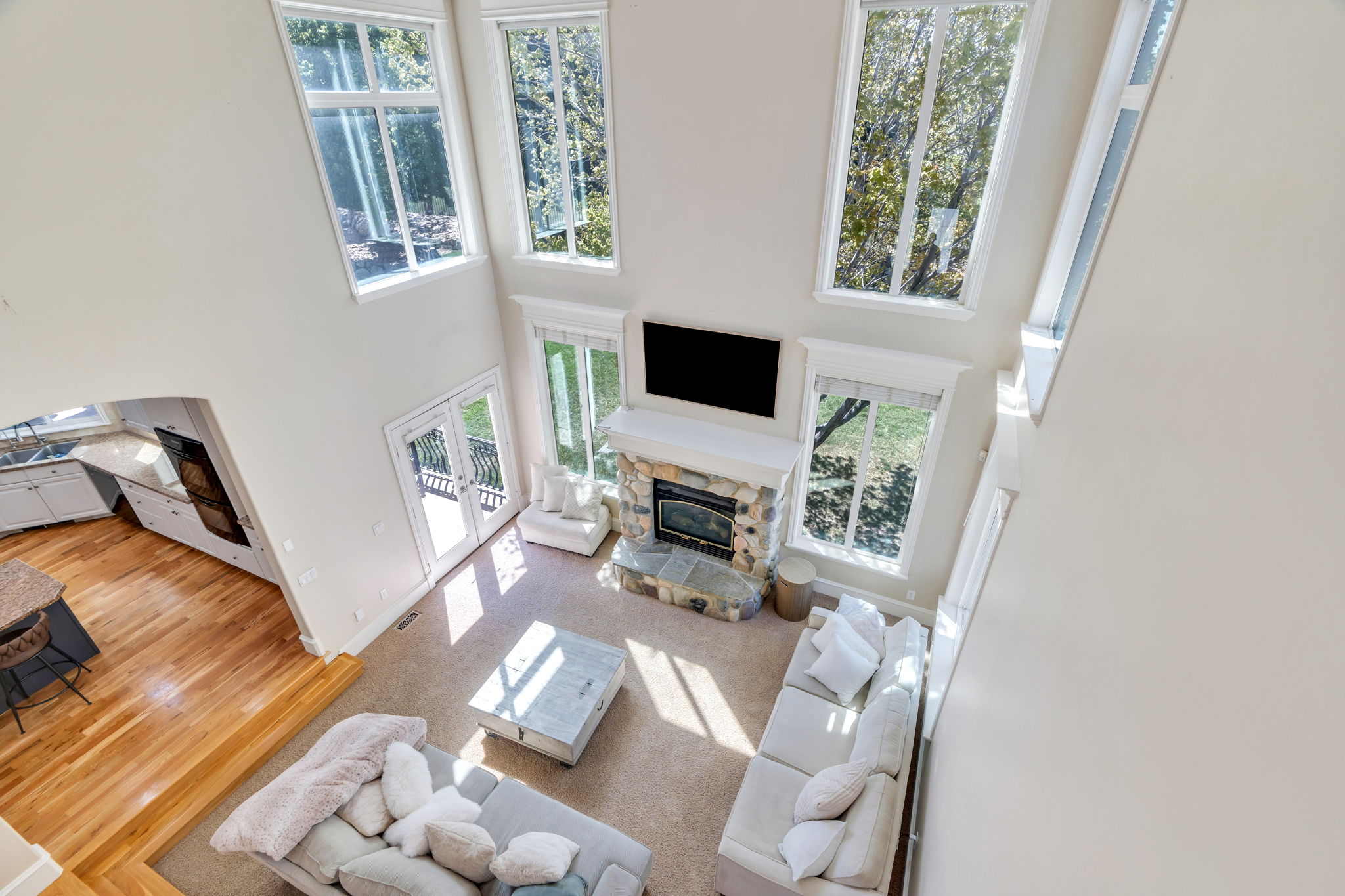 Two-story living area featuring a stone fireplace, large windows, French doors, carpet flooring, and an open concept to the kitchen with wood-finish flooring