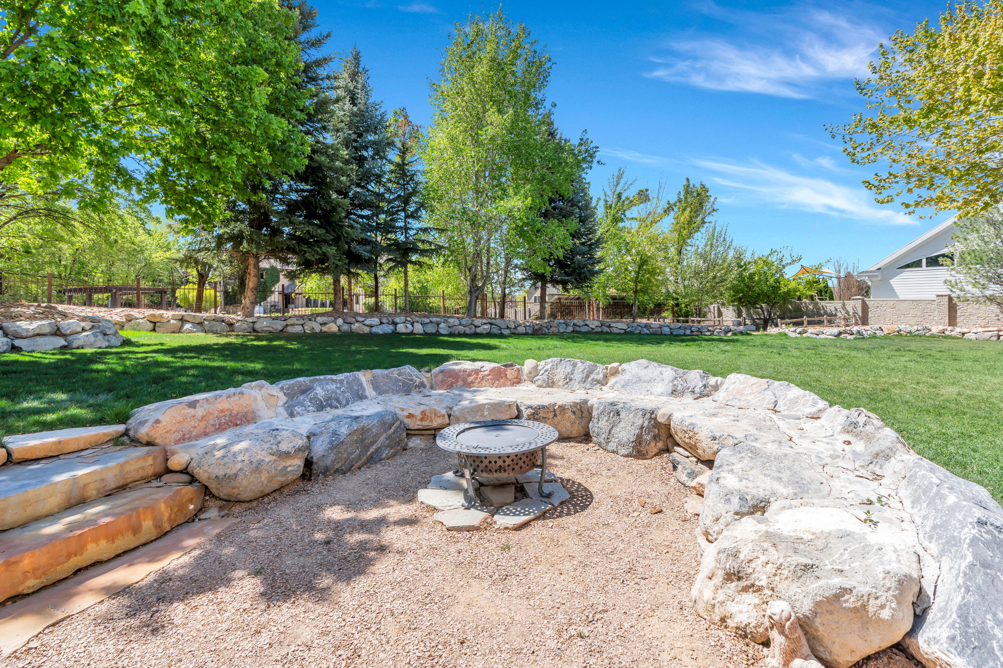 Expansive exterior space featuring a stone-lined fire pit area with gravel ground cover, bordered by a substantial natural rock retaining wall