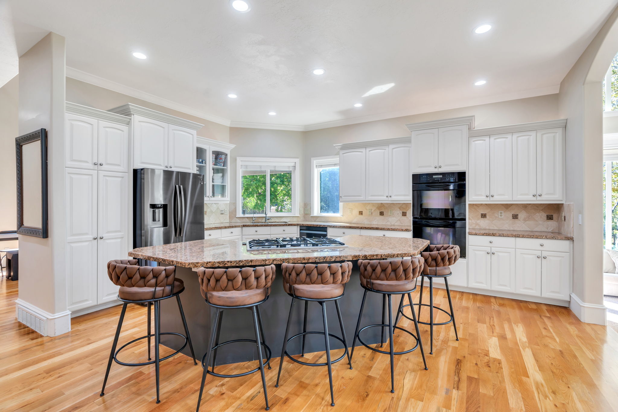 Open-concept kitchen featuring an expansive center island with a built-in cooktop, light wood-finish flooring, white cabinetry, integrated stainless steel refrigerator, and double wall ovens