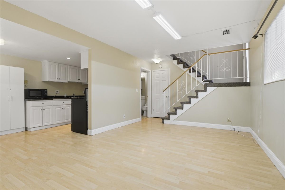 Open-concept living space featuring light wood-finish flooring, a contemporary staircase with ornamental railing, a compact kitchen with white cabinetry and dark countertops, recessed lighting, and a window with blinds