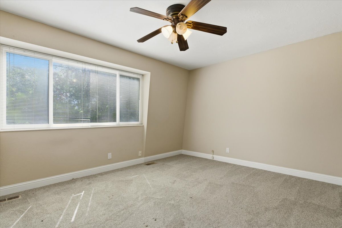 Bright interior space featuring a multi-pane window with horizontal blinds, neutral-toned walls, gray textured carpet, white baseboards, and a ceiling fan with integrated lighting