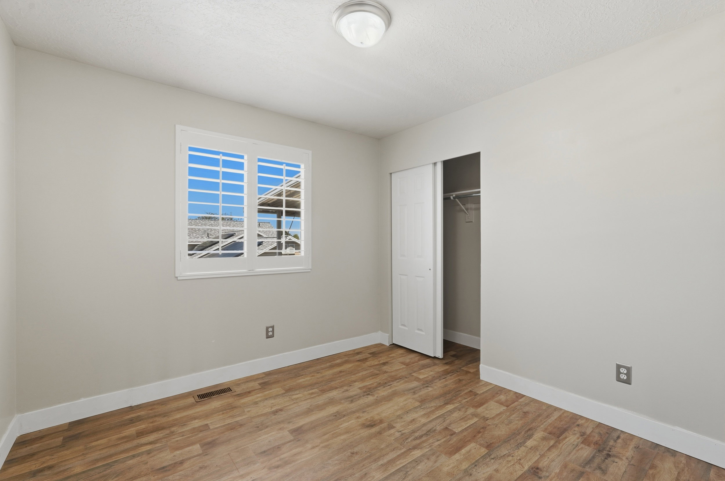 Neutral-toned room featuring a window with white shutters, wood-finish flooring, a built-in closet with bi-fold doors, and a ceiling-mounted light fixture