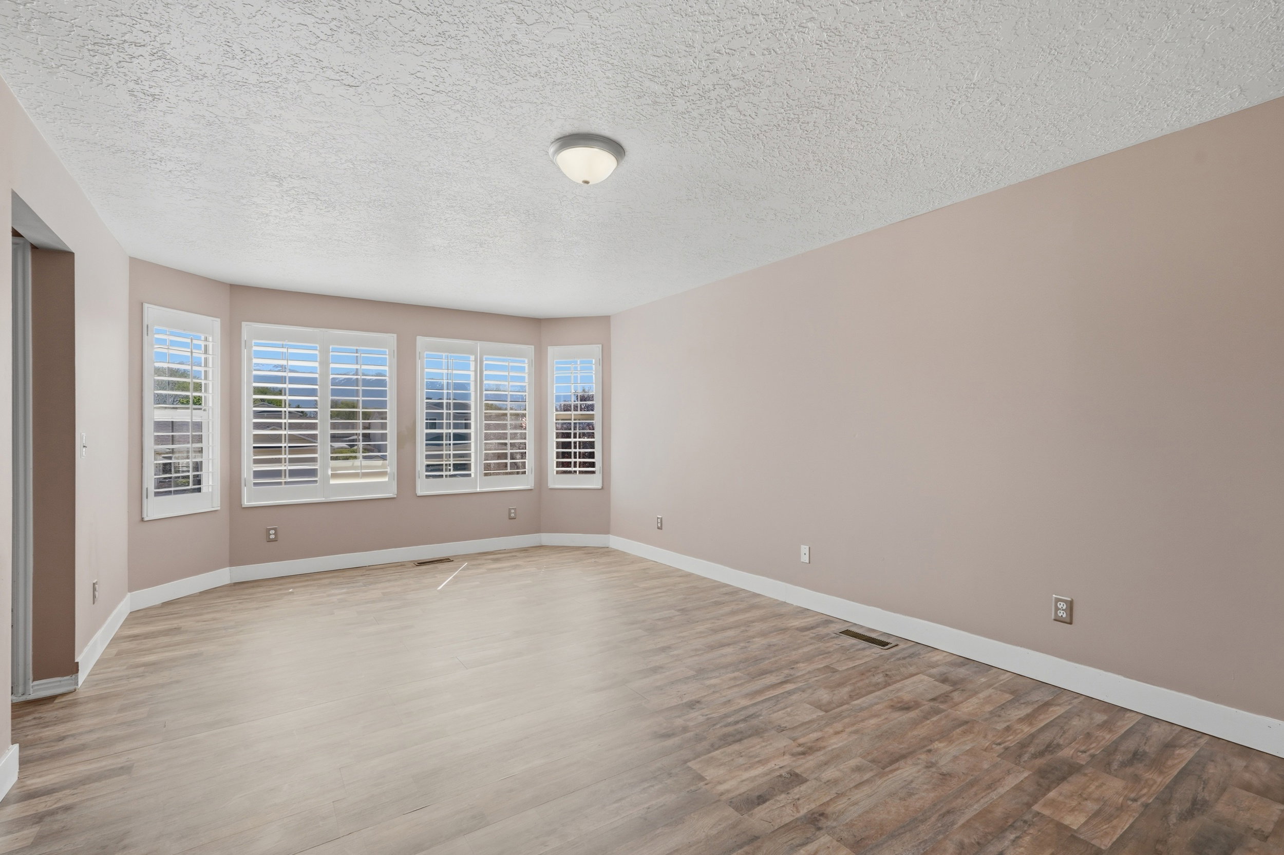 Expansive room featuring multiple windows with plantation shutters, wood-finish flooring, neutral wall tones, white baseboards, and a flush-mount ceiling light fixture