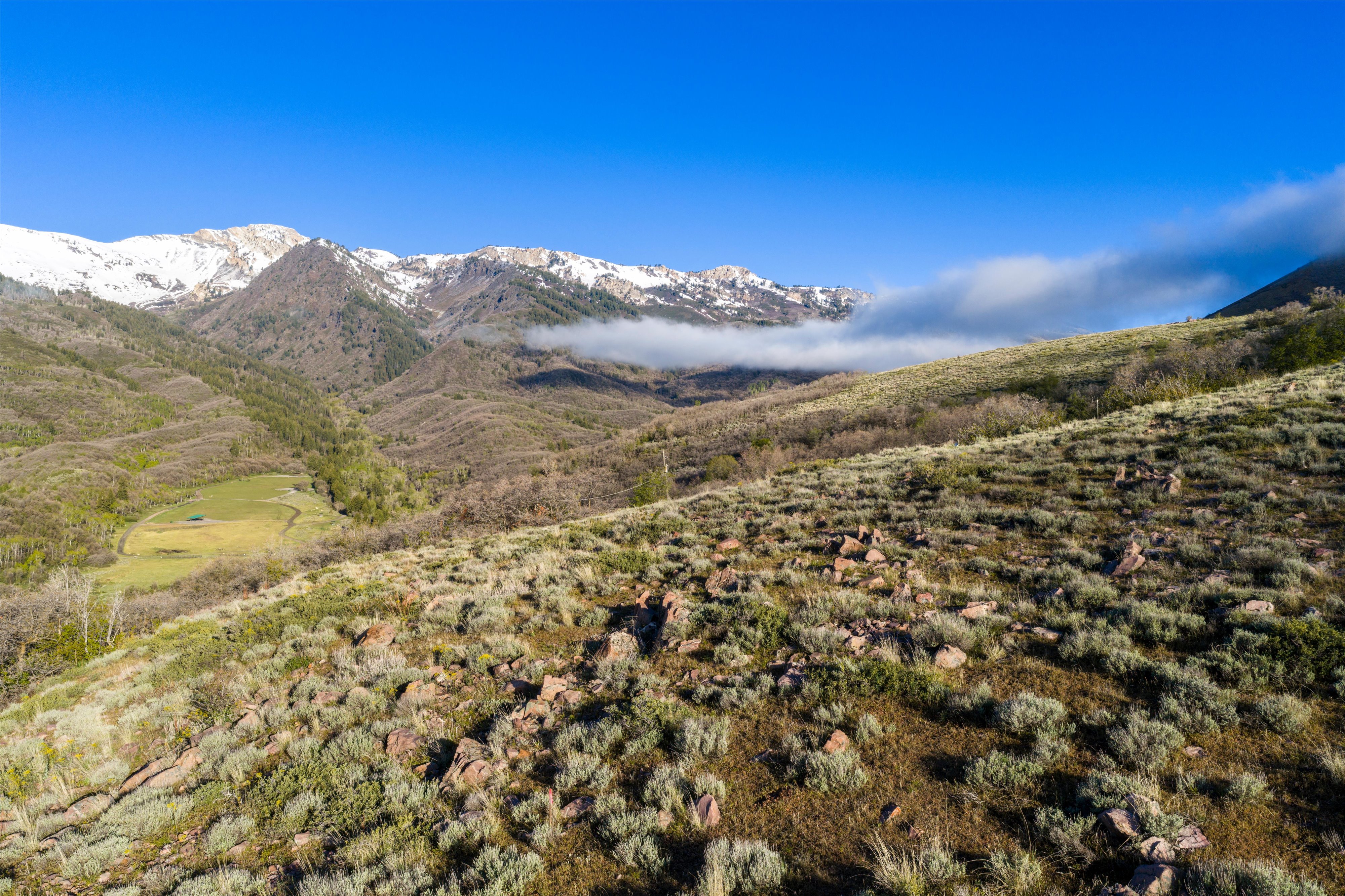 Expansive mountain landscape featuring varied terrain, a clear blue sky, and distant snow-capped peaks