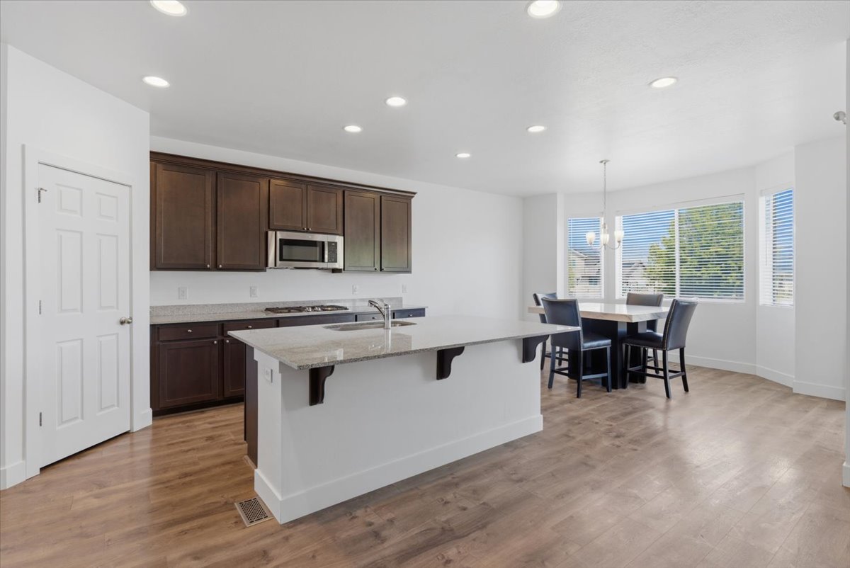 Open-concept kitchen featuring dark wood cabinetry, light granite countertops, stainless steel appliances, a center island with sink, and wood-finish flooring extending to the dining area with bay windows