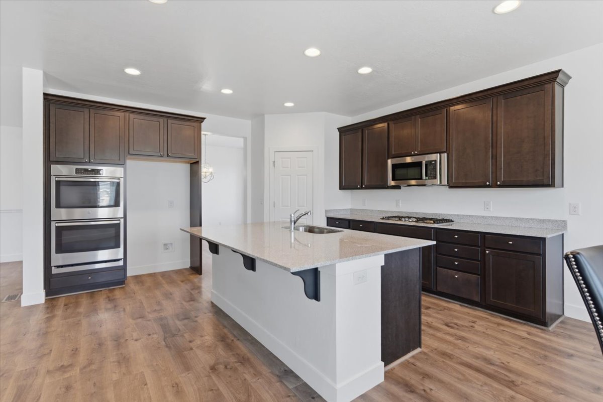 Spacious kitchen featuring an expansive center island with an undermount sink, dark wood-finish cabinetry, stainless steel double wall ovens, a built-in microwave, and wood-finish flooring