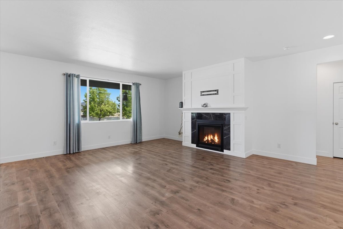 Spacious room featuring wood-finish flooring, a contemporary fireplace with a black stone surround and white mantel, a large window, recessed lighting, and white baseboards