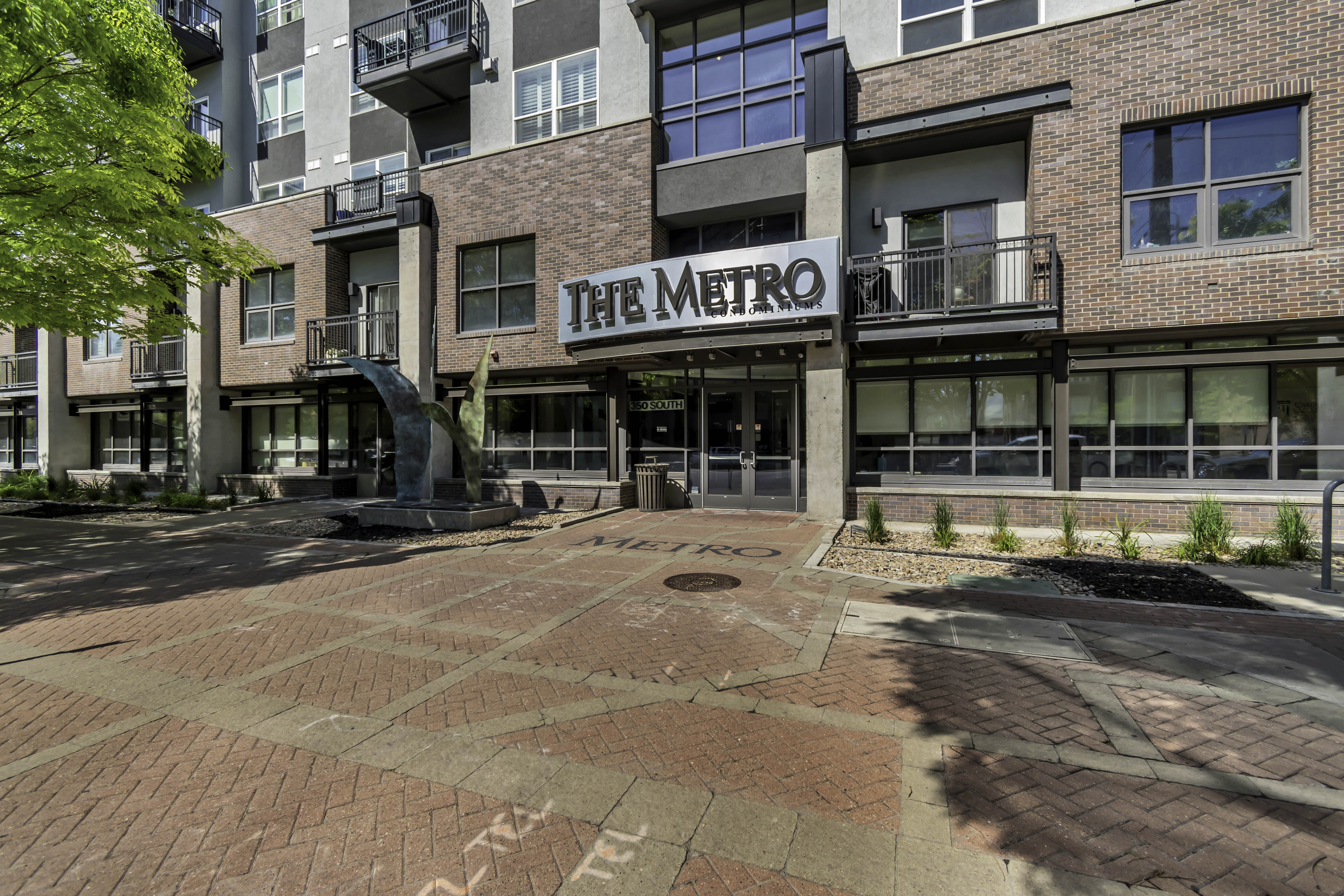 Modern multi-story building featuring brick and paneling exterior, ground-level entry with "THE METRO" signage, multiple balconies with metal railings, large storefront windows, and a paver walkway with a prominent art installation