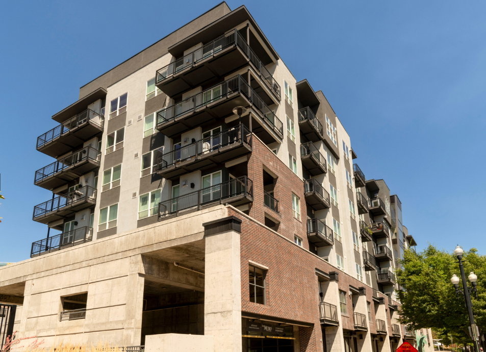 Modern multi-story building featuring individual balconies with black railings, a light-colored facade with dark gray accents, and lower-level brick detailing