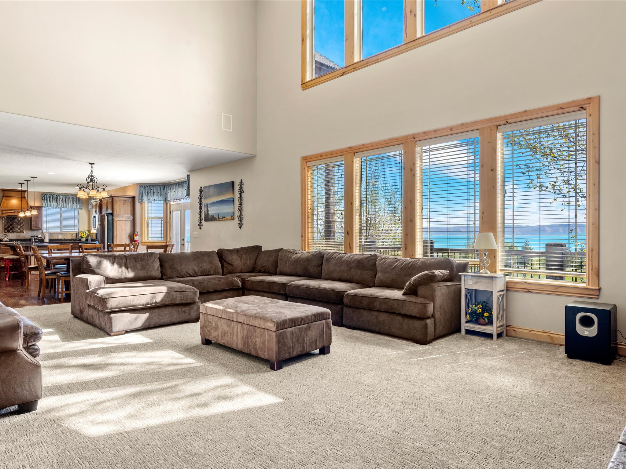 Spacious living room featuring high ceilings, large windows with natural wood trim, light-colored carpet, and an open-concept design extending to the kitchen area