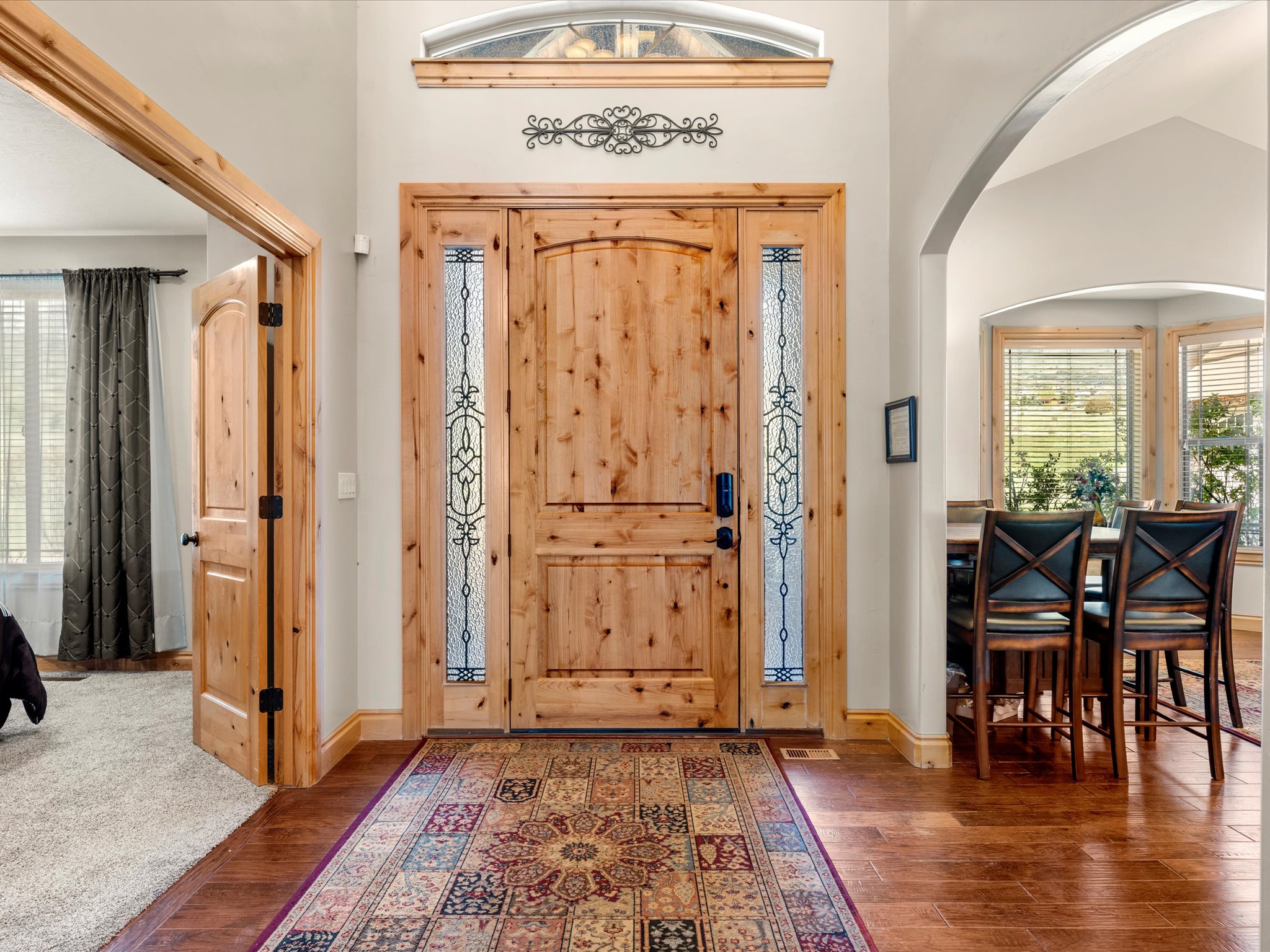 Formal living room on right, main floor bedroom on left .  Natural wood entry door with decorative glass sidelights, transom window, wood-finish flooring, and arched entryway