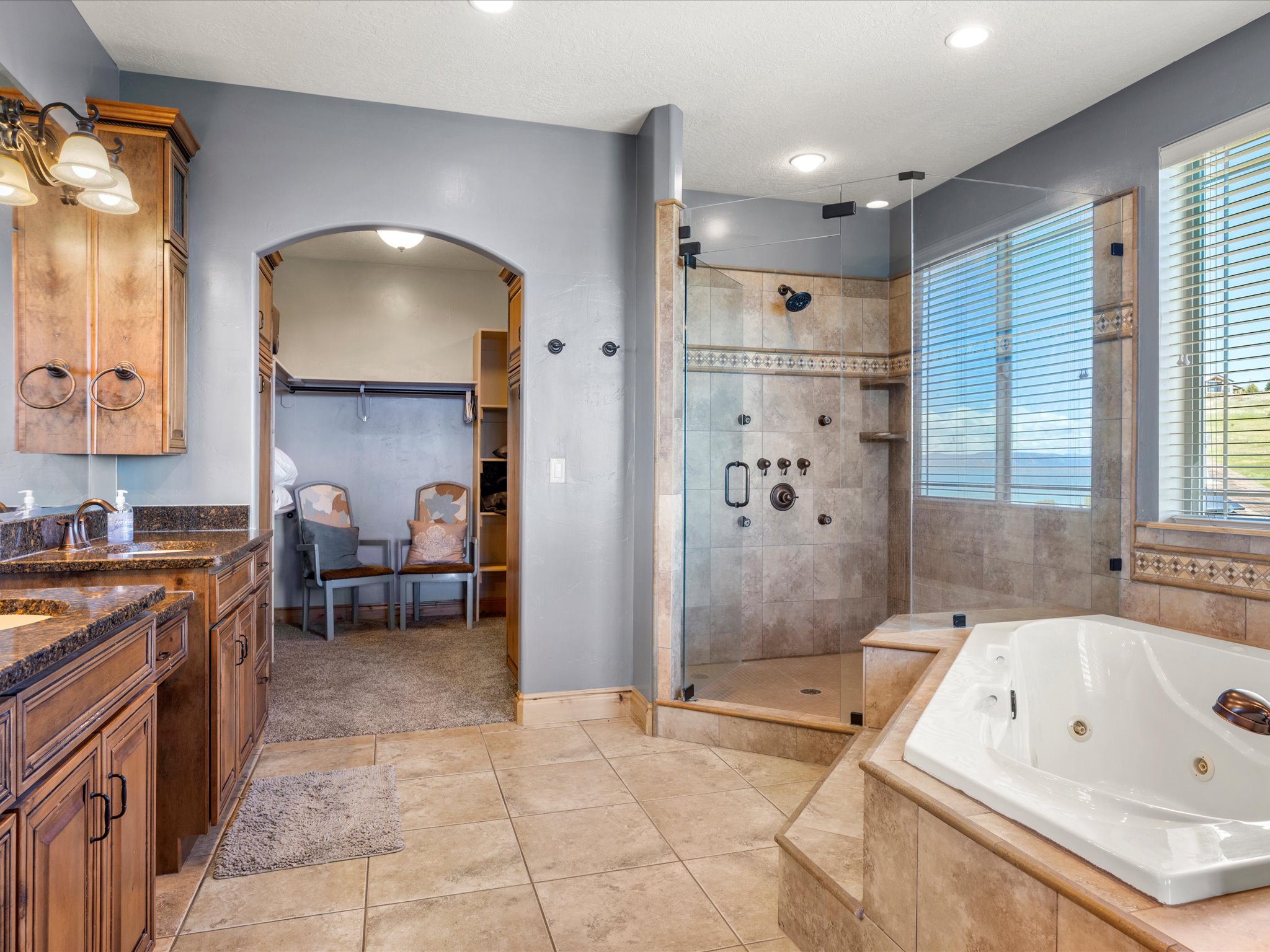 Spacious bathroom featuring a jetted soaking tub, a tiled walk-in shower with multiple showerheads, dual vanity with granite countertops, and an arched entry to a separate walk-in closet