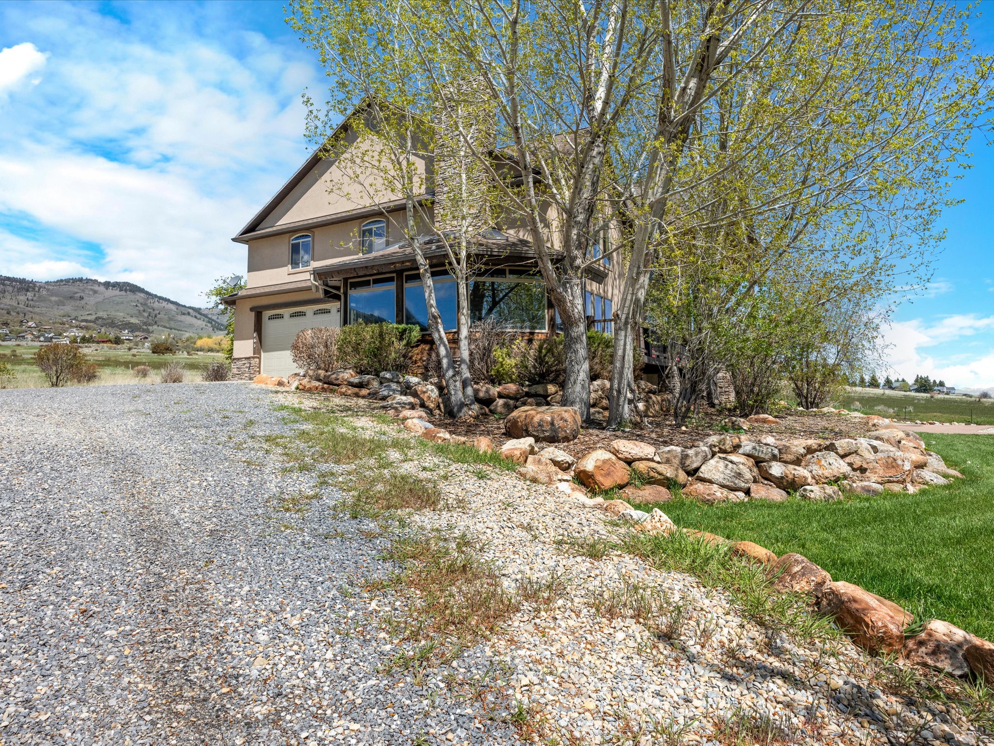 Stucco exterior residence featuring a two-car garage, multiple large windows, mature trees, and a stone retaining wall