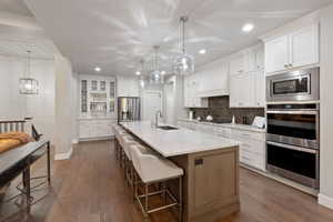 Contemporary kitchen featuring wood-finish flooring, white perimeter cabinetry, stainless steel appliances, a spacious central island with a light-toned countertop, and pendant lighting fixtures