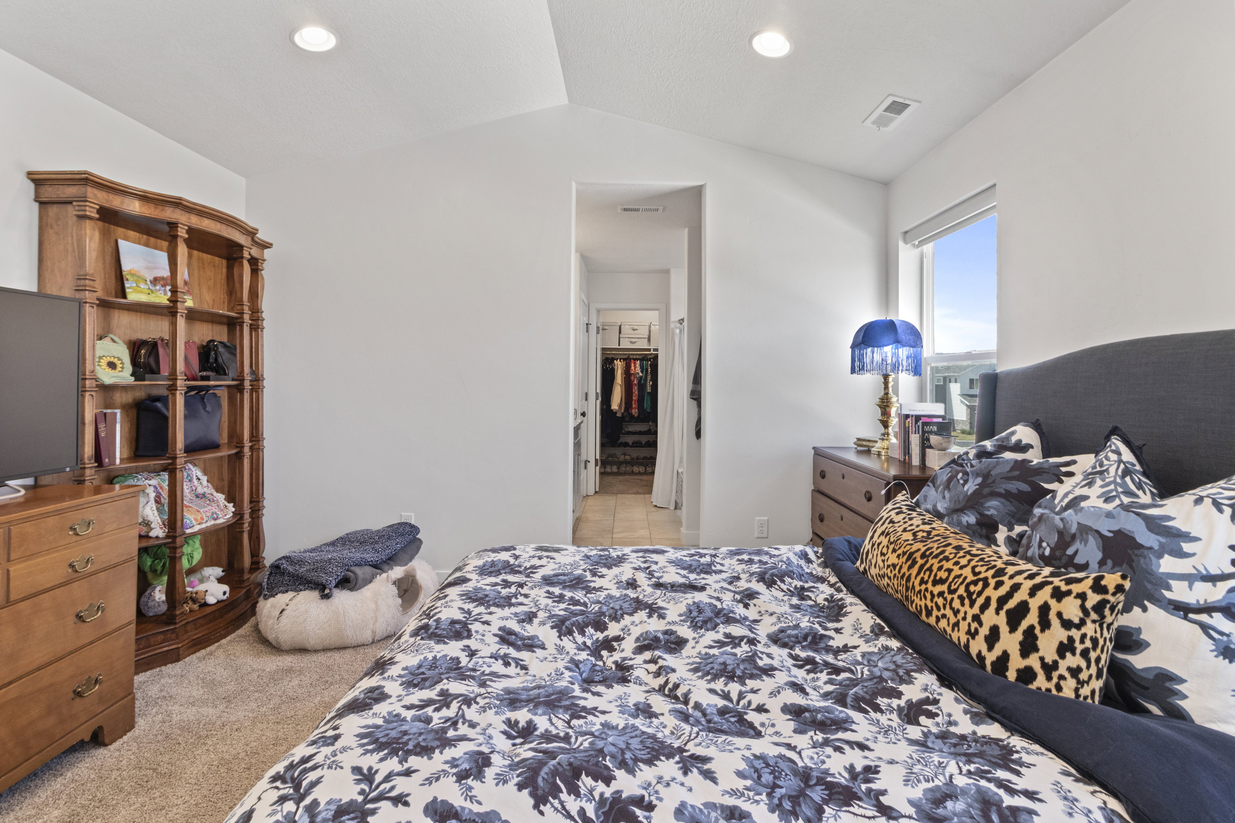 Bright bedroom featuring vaulted ceilings, recessed lighting, carpet flooring, a window with outdoor scenery, and an open doorway to an interior space with tile flooring